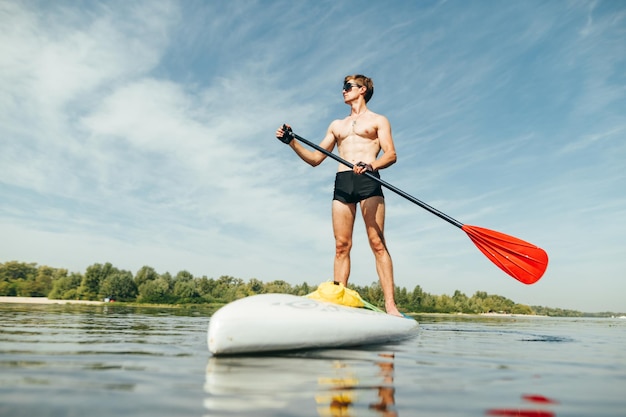 Un homme musclé athlétique avec un torse sourd se tient sur une planche de sup avec une rame à la main et regarde au loin avec un visage sérieux