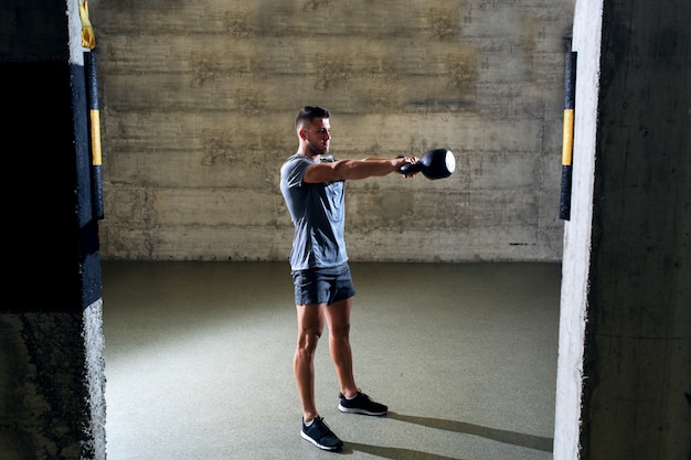 Homme musclé dédié en vêtements de sport, faire des exercices avec kettlebell en se tenant debout dans une salle de sport cross-fit.