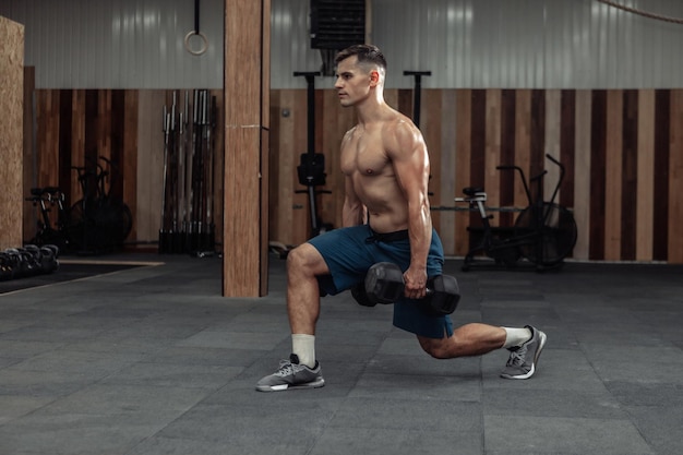 Homme musclé entraînant ses jambes, faisant des fentes avec des haltères lourds dans une salle de sport moderne. Concept de mode de vie sain