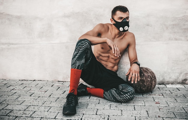 Homme musclé de remise en forme assis sur un sol en béton avec médecine-ball dans un masque en plein air à côté de la grille après les exercices Espace de copie pour la publicité Sportif torse nu faisant de l'exercice