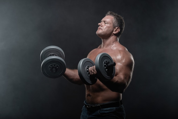 Homme musclé s&#39;entraîne avec des haltères sur fond noir en fumée