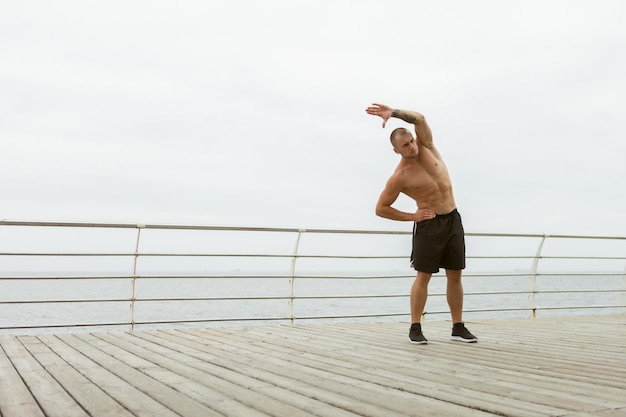 Homme musclé avec un torse nu faisant des étirements corporels avant de s'entraîner sur la plage