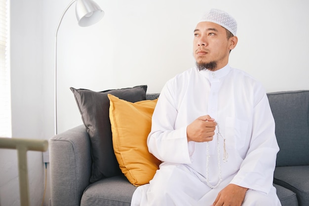 Un homme musulman asiatique assis sur un canapé prie avec des perles de prière.
