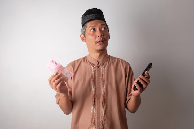 Photo homme musulman asiatique confus tenant de l'argent et un téléphone indonésiens