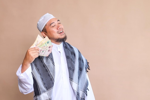 Un homme musulman asiatique montrant de l'argent en espèces et regardant de côté sur un fond beige