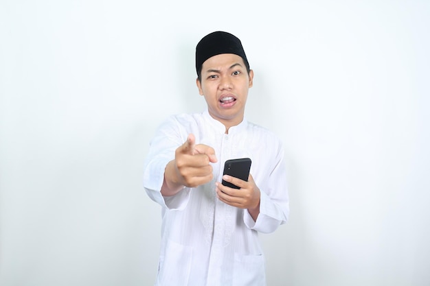 Photo un homme musulman tenant un téléphone portable pointant vers l'avant vers la caméra avec une expression confuse isolé sur un fond blanc
