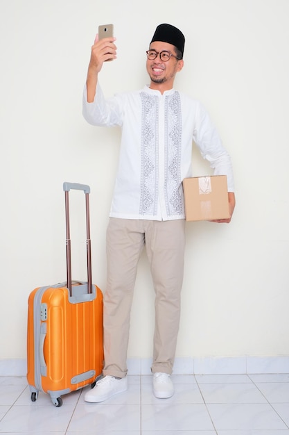 Un homme musulman avec une valise et un carton qui regarde son téléphone avec une expression heureuse.