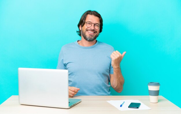 Homme néerlandais senior dans une table avec un ordinateur portable isolé sur fond bleu pointant vers le côté pour présenter un produit