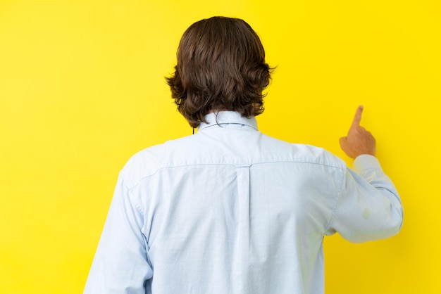 Homme néerlandais de télévendeur travaillant avec un casque isolé sur fond jaune pointant vers l'arrière avec l'index