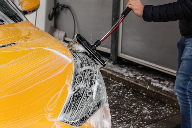 Homme nettoyant sa voiture dans un lave-auto en libre-service, brosse laissant des coups de shampoing sur le phare avant.