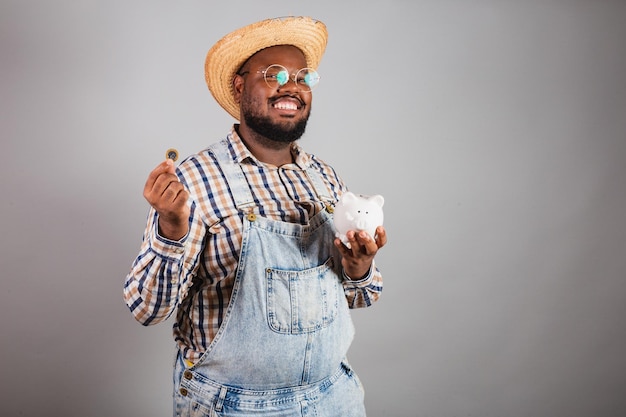 Homme noir brésilien portant des vêtements de campagne de la festa junina festa de sao joao arraia tenant une tirelire et une économie de la finance monétaire