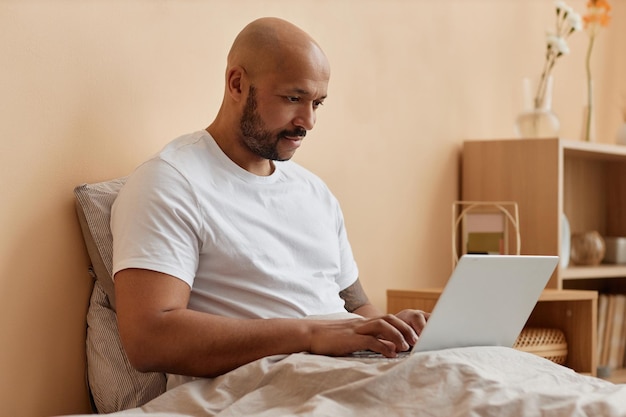 Un homme noir chauve qui utilise un ordinateur portable dans son lit à la maison.