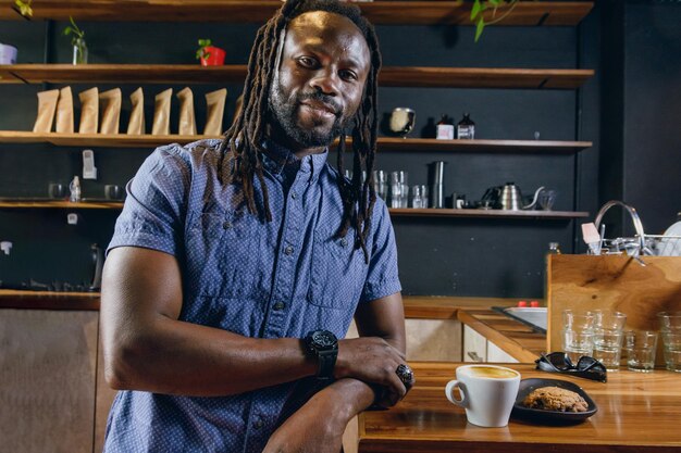 Photo homme noir avec des dreadlocks et une barbe mangeant du café avec des biscuits heureux souriant en regardant la caméra debout à côté du comptoir à l'intérieur de l'espace de copie du restaurant