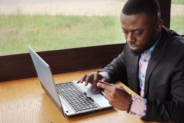 L'homme noir effectue le paiement par carte bancaire sur un ordinateur portable. Achats en ligne assis dans un café. Un jeune homme d'affaires afro-américain achète en ligne en saisissant ses données sur l'ordinateur à partir d'une carte de crédit, vue latérale. Client de type.