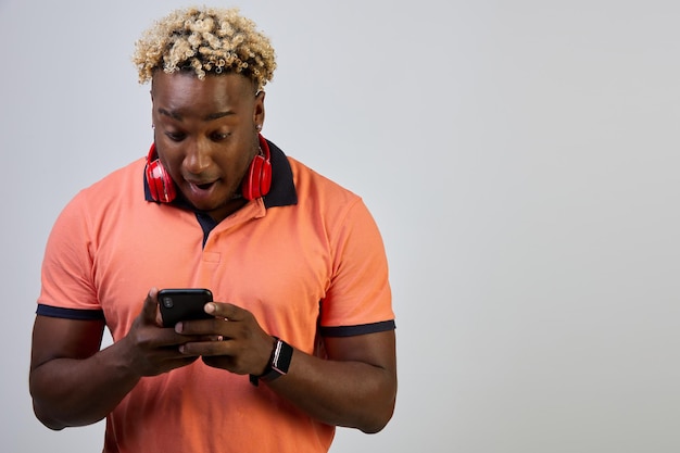 Un homme noir émotionnel avec un casque autour du cou utilise un smartphone debout sur un fond blanc