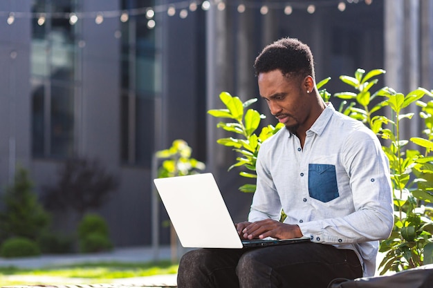 Homme noir indépendant travaillant en ligne à l'aide d'un ordinateur portable assis sur un banc à l'extérieur du bâtiment moderne du bureau dans le parc urbain de la ville dans la rue. Heureux étudiant afro-américain en plein air décontracté en tapant sur le clavier