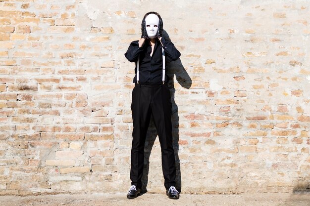 Photo un homme en noir avec un masque blanc sur le visage contre un mur de briques.