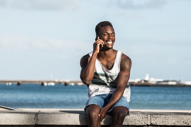 Homme noir positif parlant sur smartphone au bord de mer en été