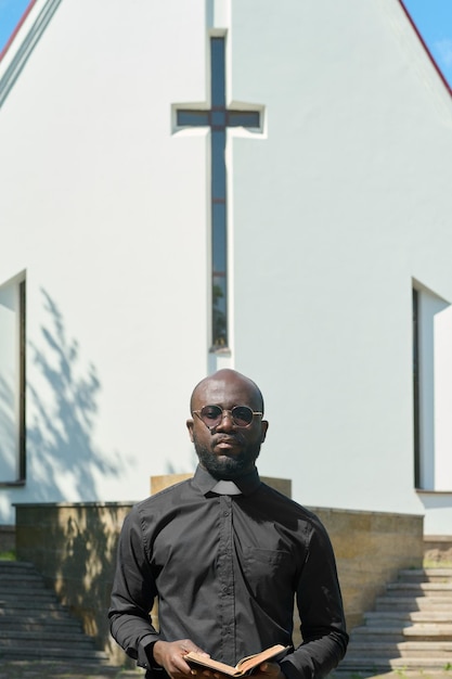 Homme noir sérieux avec un évangile ouvert dans les mains regardant la caméra contre l'église