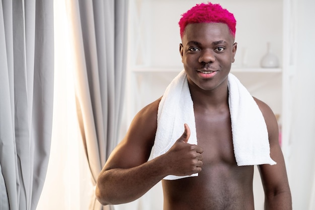 Homme noir sexy Routine du matin Bon choix Soins de traitement Homme africain torse nu métrosexuel souriant avec une serviette de cheveux rose vif sur l'épaule pouce vers le haut dans l'espace de copie intérieur de la salle de bain