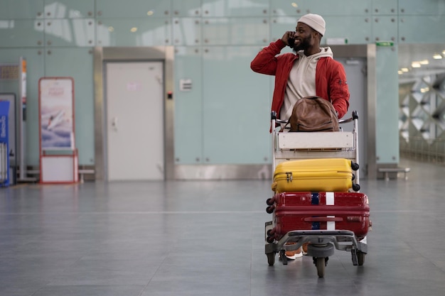 Homme noir souriant poussant un chariot à bagages marchant après son arrivée à l'aéroport parlant sur un téléphone portable