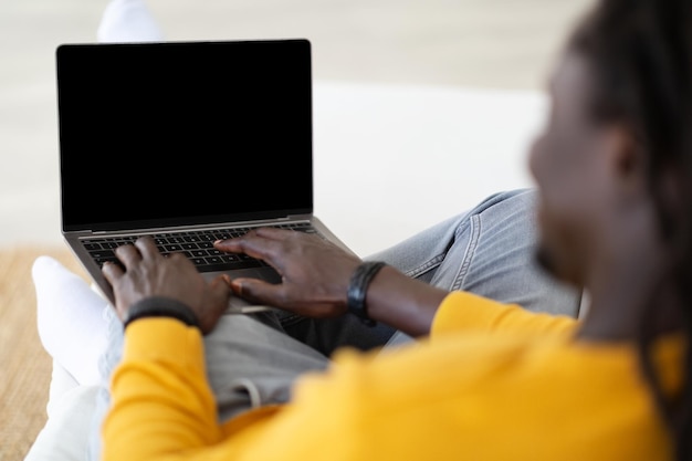 Homme noir tapant sur un ordinateur portable avec écran vide tout en étant assis sur un canapé