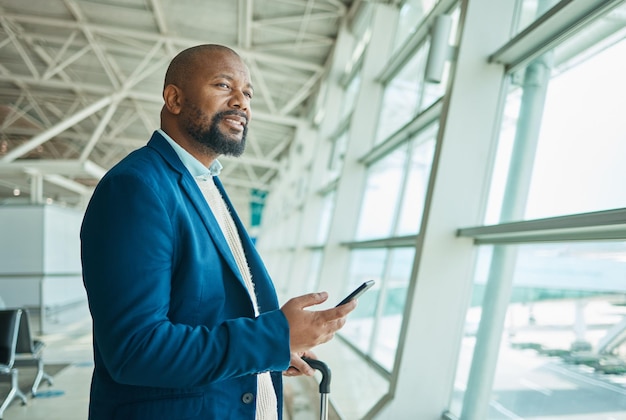 Homme noir téléphone et pensant à la fenêtre de l'aéroport pour un voyage d'affaires ou une communication en attente de vol Homme afro-américain avec le sourire en contemplant l'horaire ou les heures simples sur smartphone