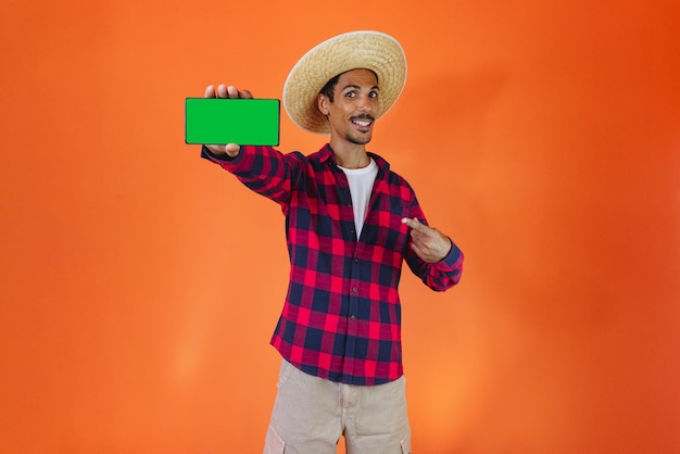 Homme noir avec une tenue de fête Junina tenant une tablette ou un téléphone portable isolé sur fond orange Jeune homme portant des vêtements traditionnels pour le festival brésilien de juin Festa Junina