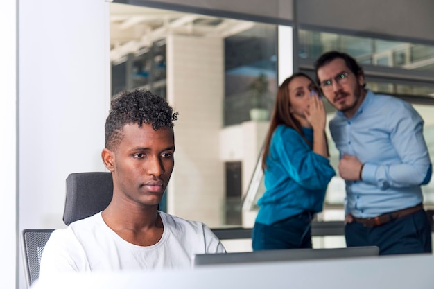 Un homme noir travaille sur un bureau et il y a deux collègues qui bavardent derrière lui