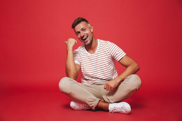 homme non rasé des années 30 en t-shirt rayé souriant, assis sur le sol avec les jambes croisées isolées sur le rouge