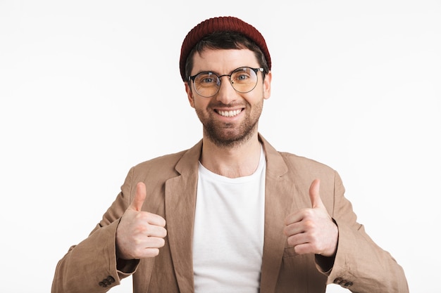 homme non rasé avec du chaume portant un chapeau et des lunettes montrant les pouces vers le haut isolé sur un mur blanc