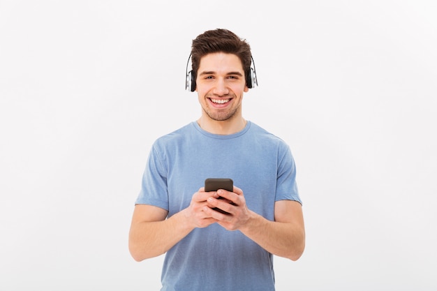 Homme non rasé en t-shirt décontracté, écouter de la musique via des écouteurs sans fil à l'aide de téléphone portable, isolé sur mur blanc