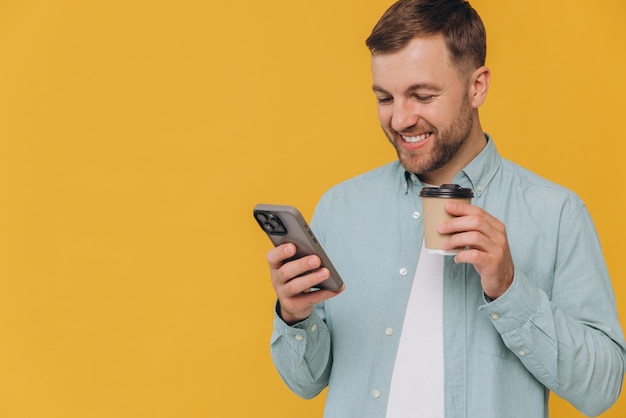 Un homme non rasé tenant un smartphone dans une main et une tasse de café à emporter dans l'autre regardant l'écran du téléphone isolé sur un fond jaune