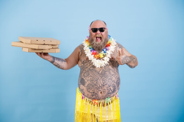 Un homme obèse joyeux avec une guirlande de fleurs tient des boîtes de pizza sur fond bleu clair