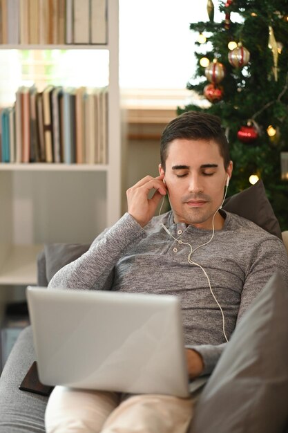 Homme occasionnel s'asseyant sur le sofa près de l'arbre de Noël et à l'aide de l'ordinateur portable