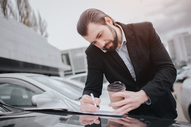 Un homme occupé est pressé, il n'a pas le temps, il va parler au téléphone en déplacement. Homme d'affaires faisant de multiples tâches La vente de voitures, l'acheteur ou le vendeur consiste à remplir des formulaires vierges sur la voiture.