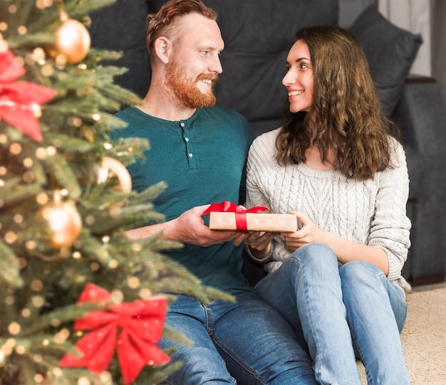 Un homme offre un cadeau à une femme assis près du sapin de Noël