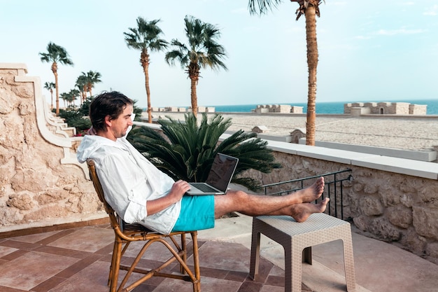 Un homme avec un ordinateur portable dans les mains se repose et travaille comme indépendant sur fond de palmiers