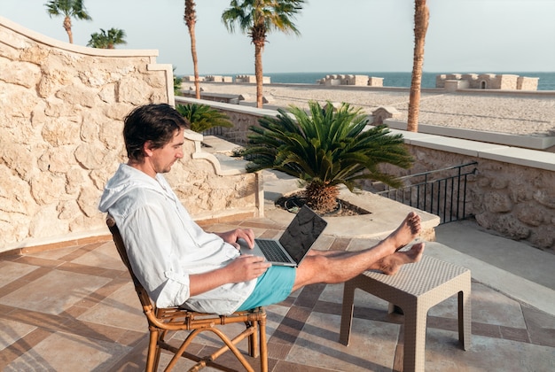 Un homme avec un ordinateur portable dans ses mains se repose et travaille comme pigiste sur la table des palmiers