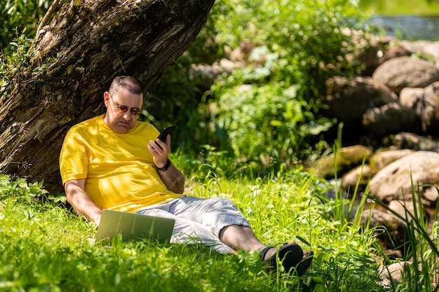 Homme avec ordinateur portable et smartphone travaillant à l'extérieur dans le pré au bord de la rivière concept de bureau en plein air