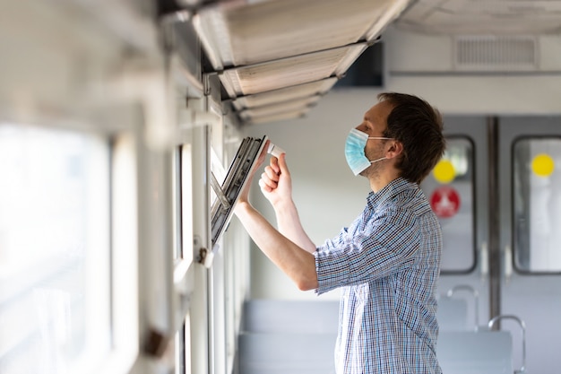 Un homme ouvre une fenêtre en train pour respirer l'air frais et la ventilation, portant un masque de protection lors d'un nouveau changement normal après l'épidémie de Covid-19