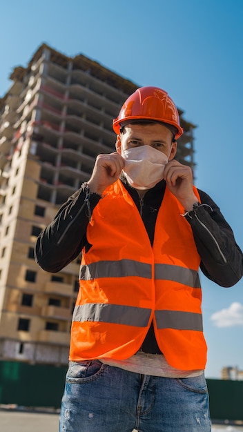 Homme ouvrier du bâtiment en salopette mettant un masque médical sur le visage sur fond de maison en construction