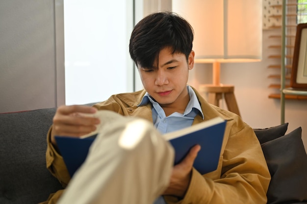 Homme paisible du millénaire lisant un livre intéressant sur un canapé confortable profitant d'un week-end de loisirs à la maison