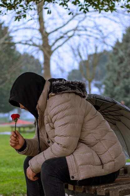 Un homme avec un parapluie est assis sous un arbre tient une fleur rouge sur un banc de parc jour de printemps pluvieux