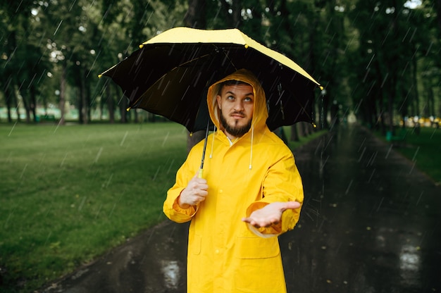 Homme avec parapluie marchant dans le parc d'été en jour de pluie. Personne de sexe masculin en cape de pluie et bottes en caoutchouc, temps humide