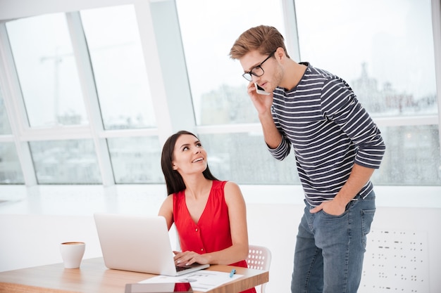 Homme parlant au téléphone et debout près de la femme près de la table au bureau près de la fenêtre