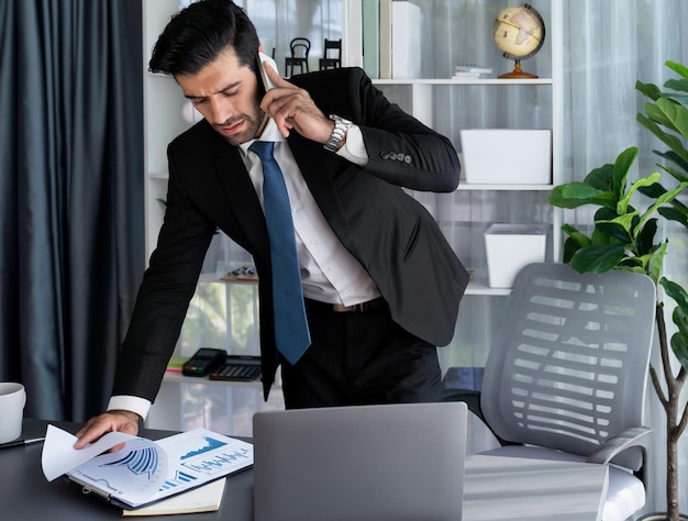 Un homme parle au téléphone tout en se tenant devant un bureau avec un ordinateur portable.