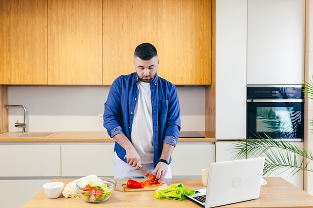 un homme passe une journée à la maison prépare un petit-déjeuner de légumes dans la cuisine un homme en vêtements de maison et avec une barbe utilise un ordinateur portable pour apprendre la cuisine en ligne