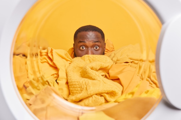 un homme à la peau foncée recouvert de tas de linge pose de l'intérieur de la machine à laver lave des vêtements sales à la maison pose