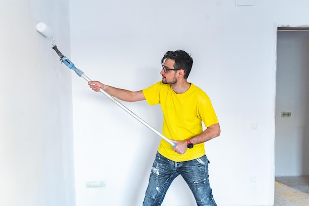 Homme peignant un mur blanc avec un long pinceau rouleau de peinture dans une maison